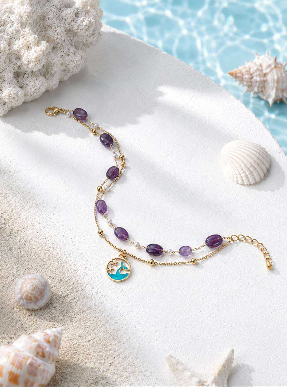 A gold-toned beaded bracelet with purple oval beads, clear spacer beads, and a circular turquoise and gold charm, styled on white sand with seashells and a blue water background.
