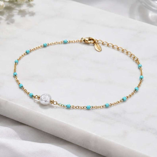 A delicate gold chain bracelet with small turquoise blue beads and a central white pearl, displayed on a white marble slab.