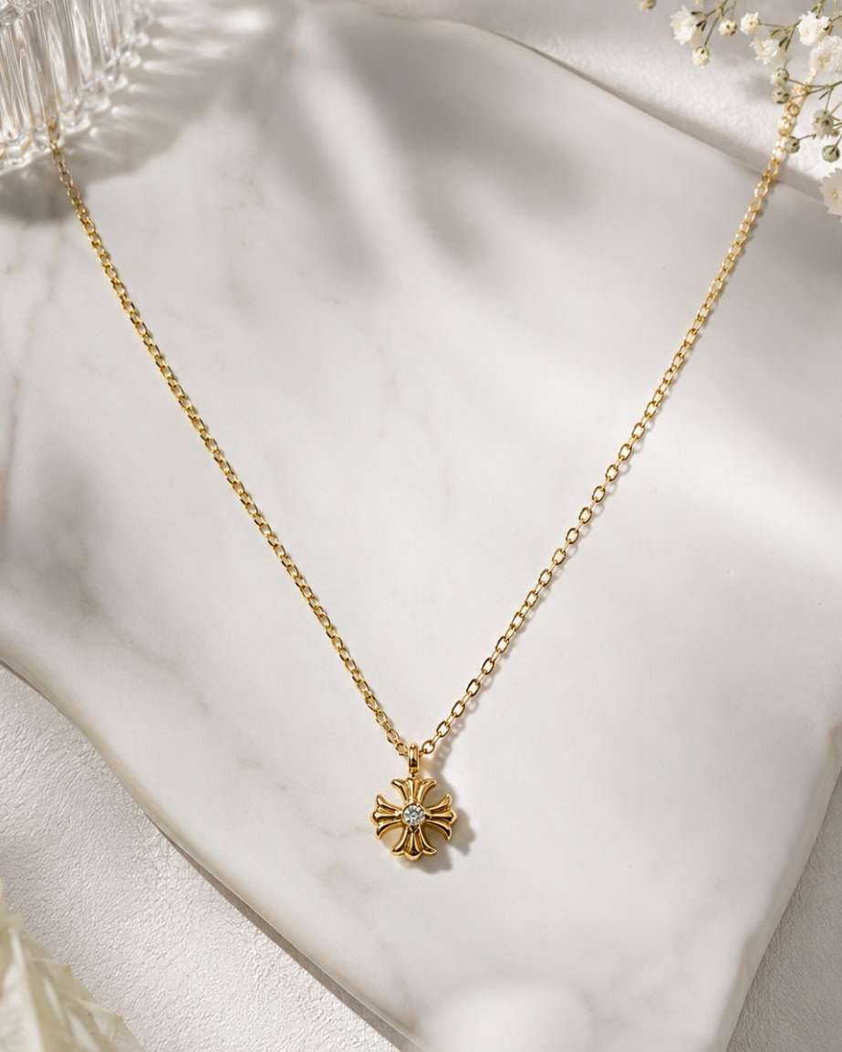 A delicate gold chain necklace with a small four-petal flower pendant, styled on draped white satin fabric with dried baby's breath flowers.