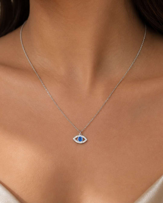 A close-up of a model's neck and chest wearing a delicate silver chain necklace with a small evil eye pendant featuring a blue center.