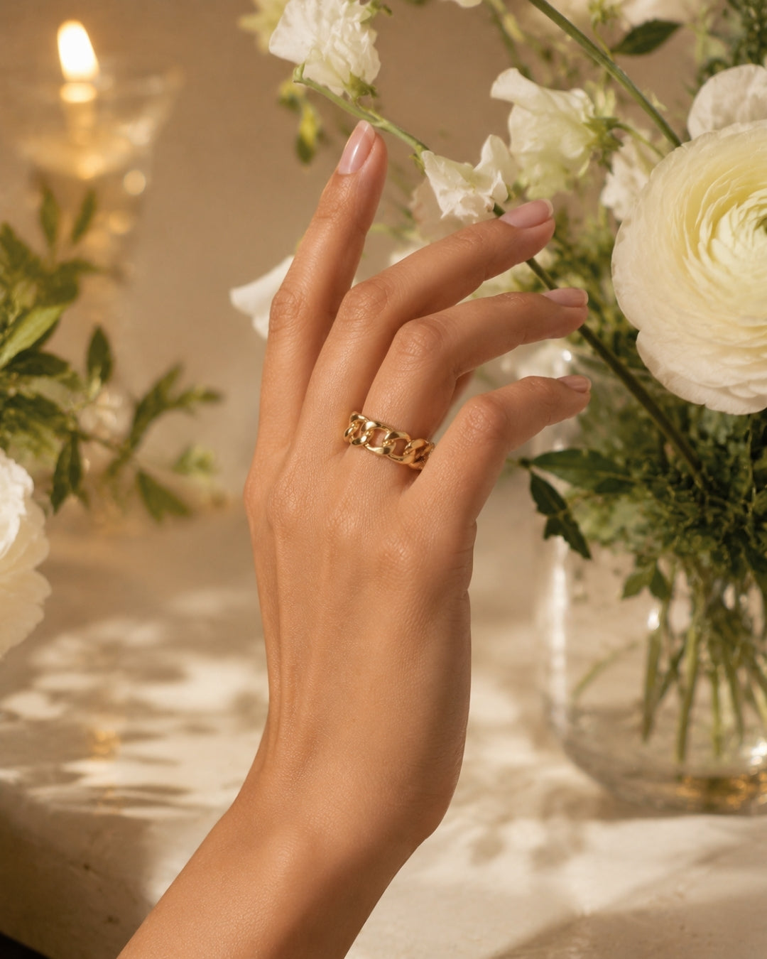 A close-up of a hand wearing a gold chain-link ring, styled with white flowers and a warm candle in the background.