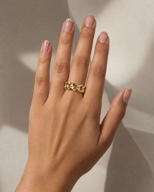 A close-up of a hand wearing a textured gold ring with a raised chain-link design, set against a warm beige background with soft shadows.
