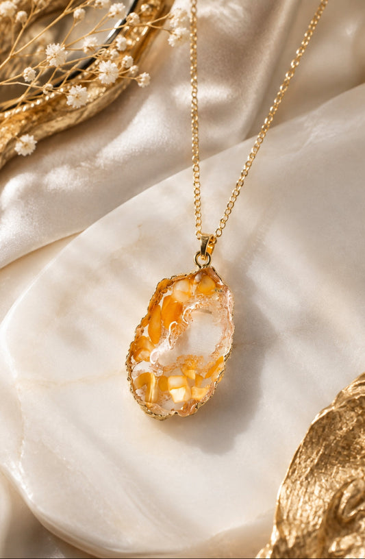 A gold chain necklace with an irregular, raw-cut geode pendant featuring orange and white crystalline formations, styled on draped cream satin fabric with dried flowers.