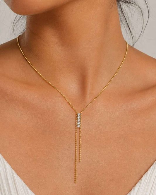 A close-up of a model wearing a delicate gold Y-necklace with a central cluster of small clear stones, paired with a white pleated top against a neutral light gray background.