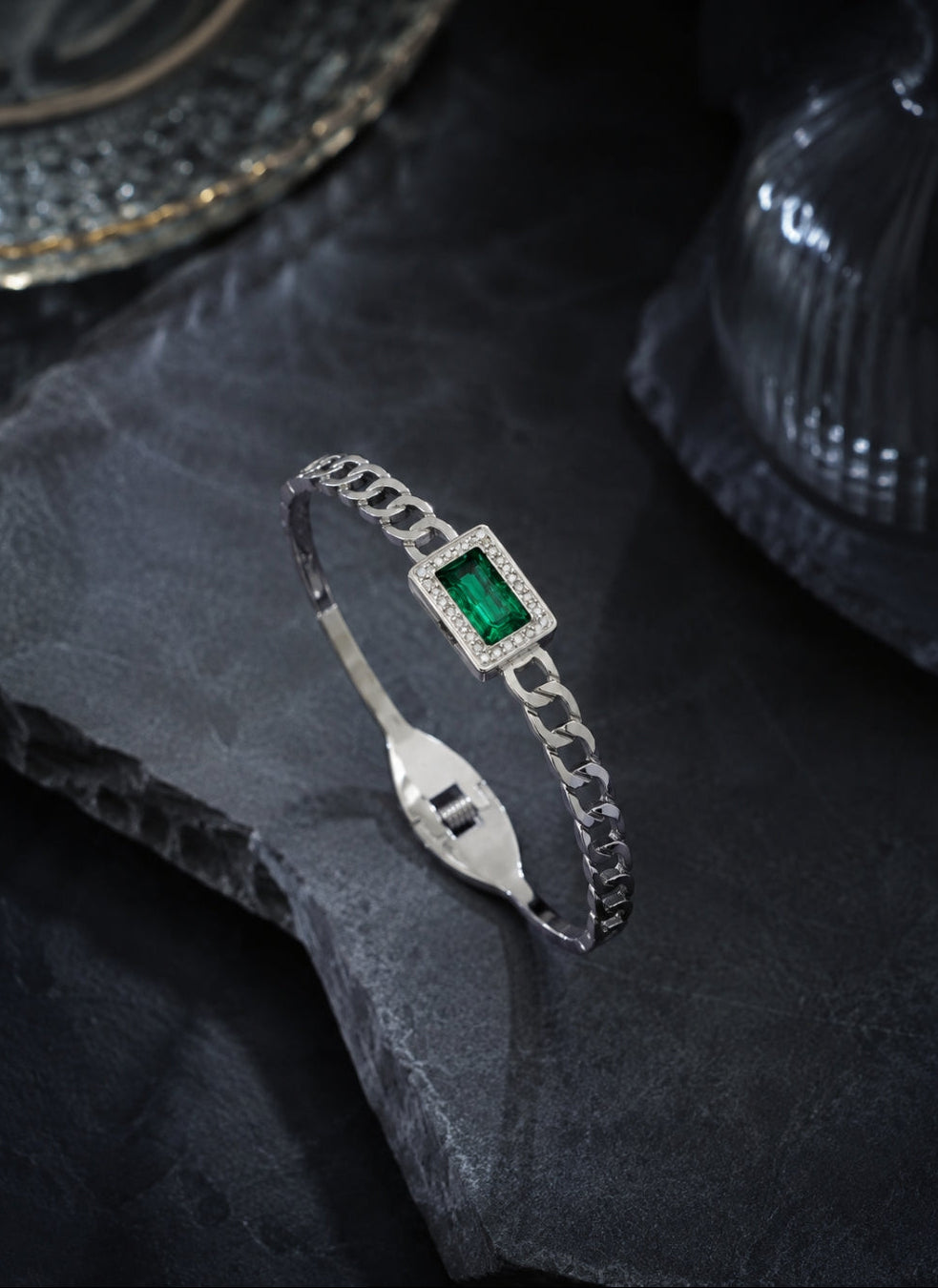 A silver chain bangle bracelet featuring a rectangular green emerald-cut center stone surrounded by small clear stones, displayed on a dark textured slate surface.