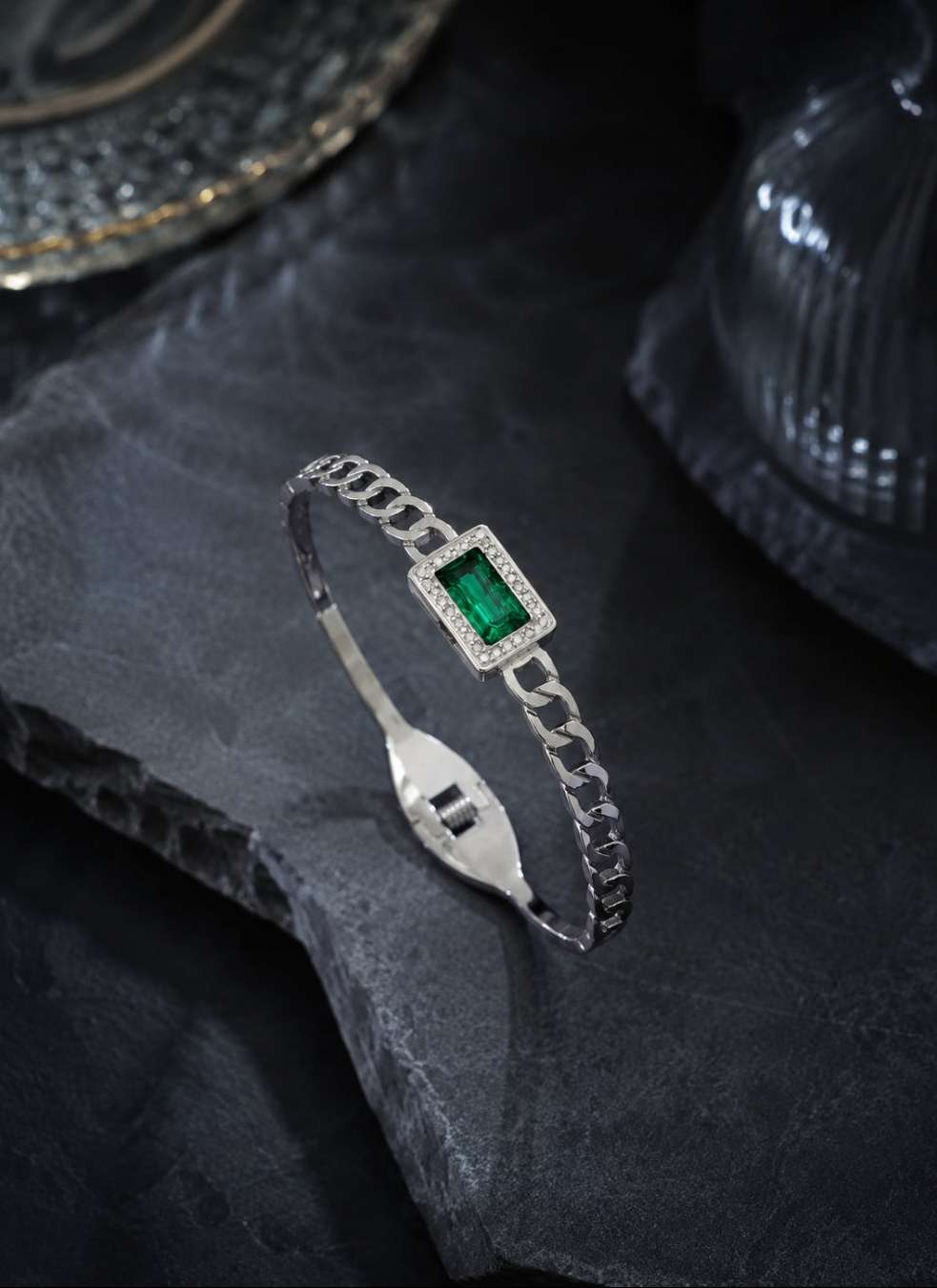 A silver chain bangle bracelet featuring a rectangular green emerald-cut center stone surrounded by small clear stones, displayed on a dark textured slate surface.