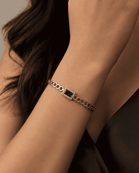 A silver chain bracelet with a central rectangular green stone, worn on a woman's wrist against a dark top and neutral background.