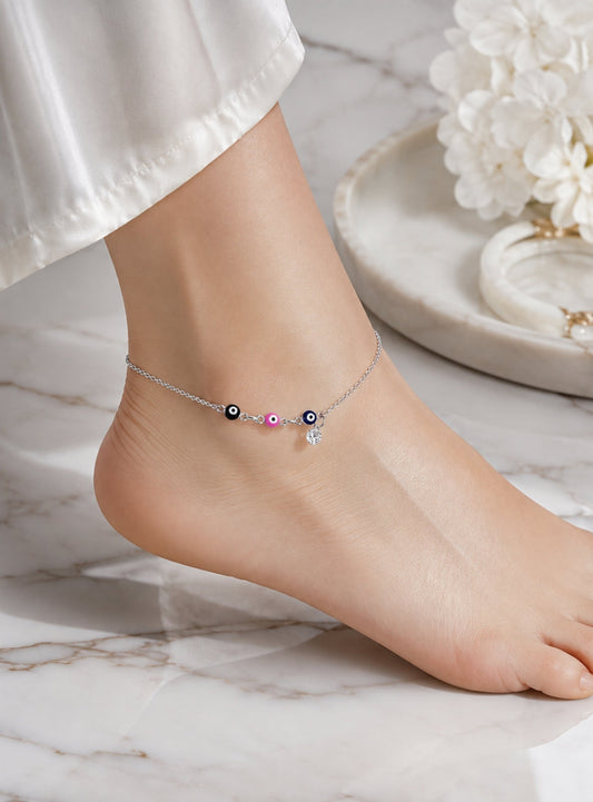 A delicate silver anklet with evil eye beads and a small dangling charm, shown worn on a person's ankle. The foot is resting on a white marble surface with white flowers in the background.
