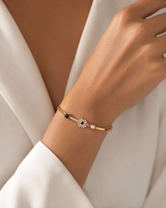 A delicate rose gold bangle bracelet with a central floral cluster of clear stones, a small white pearl, and a dark blue bead, shown worn on a woman's wrist.