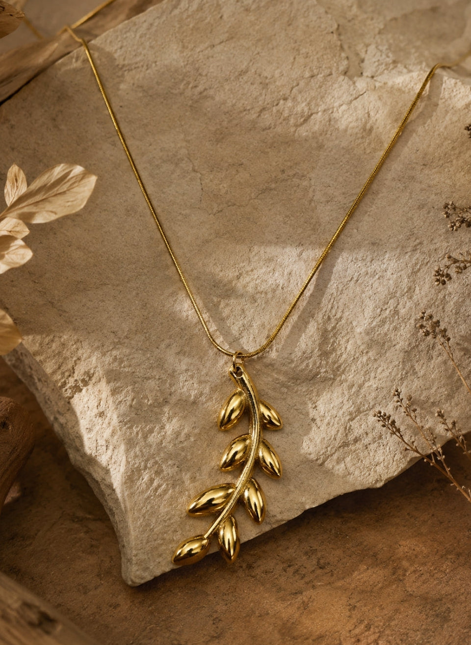 A gold leaf pendant necklace draped over a textured stone slab with dried botanical elements in the background.
