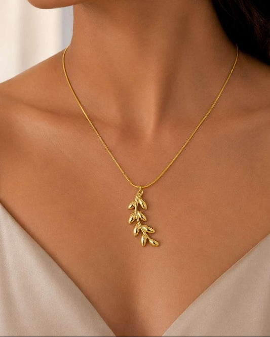 A close-up of a woman wearing a delicate gold necklace with a leafy branch pendant, styled with a beige camisole top.