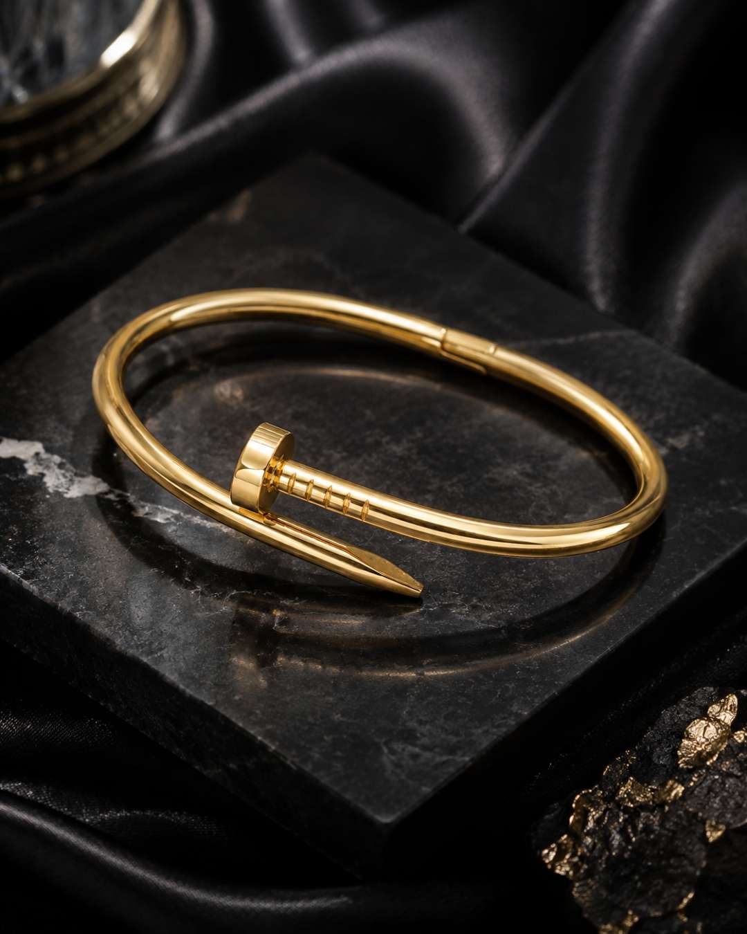A polished gold-toned metal cuff bracelet with a distinctive screw-head closure, displayed on a dark textured stone slab with black satin fabric in the background.