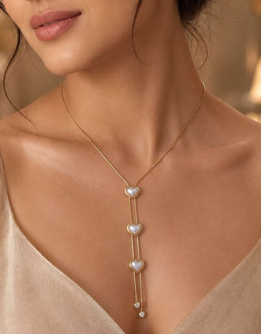 A woman wearing a delicate gold Y-necklace with three small white pearl heart charms, styled with a beige satin camisole against a warm, softly blurred background.