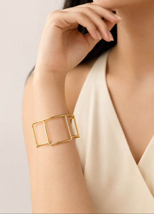 A close-up of a model's wrist wearing a wide, open-frame geometric cuff bracelet in polished yellow gold, paired with a cream-colored sleeveless top.