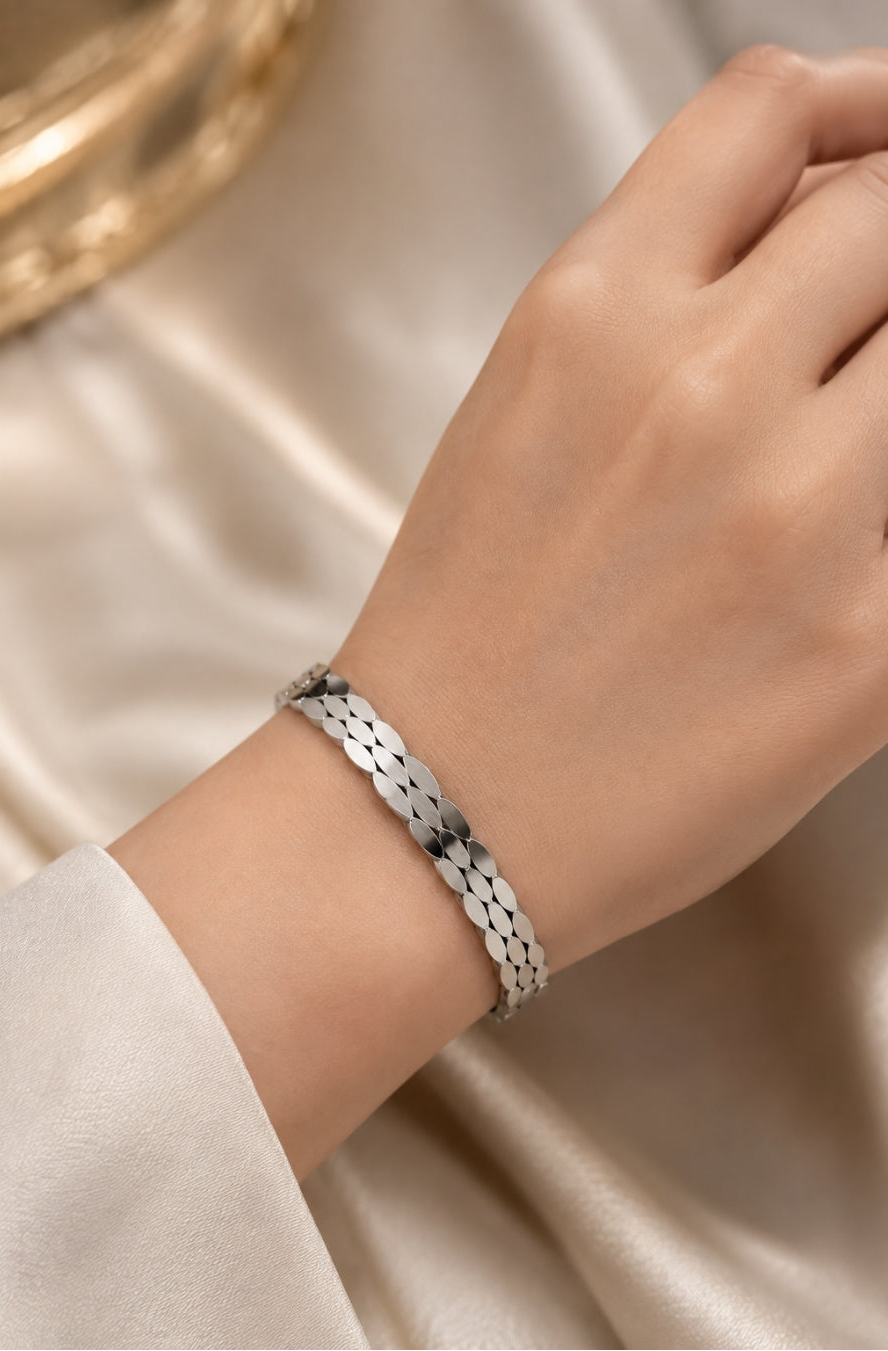 A close-up of a person's wrist wearing a silver-toned textured link bracelet, set against a soft beige satin fabric background.