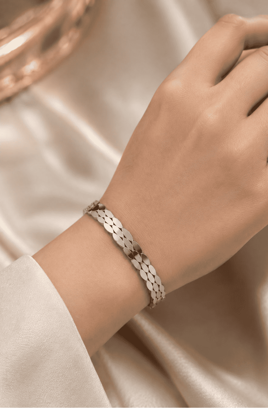 A close-up of a person's wrist wearing a silver-toned textured link bracelet, set against a soft beige satin fabric background.