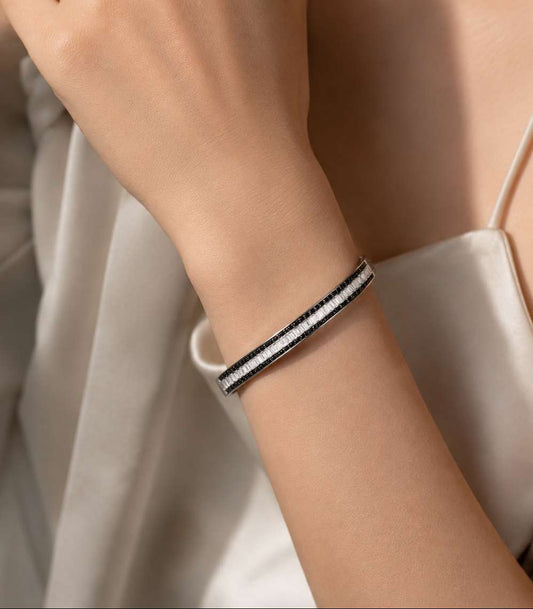 A close-up of a person's wrist wearing a slim black and white striped bangle bracelet, paired with a light-colored satin top.