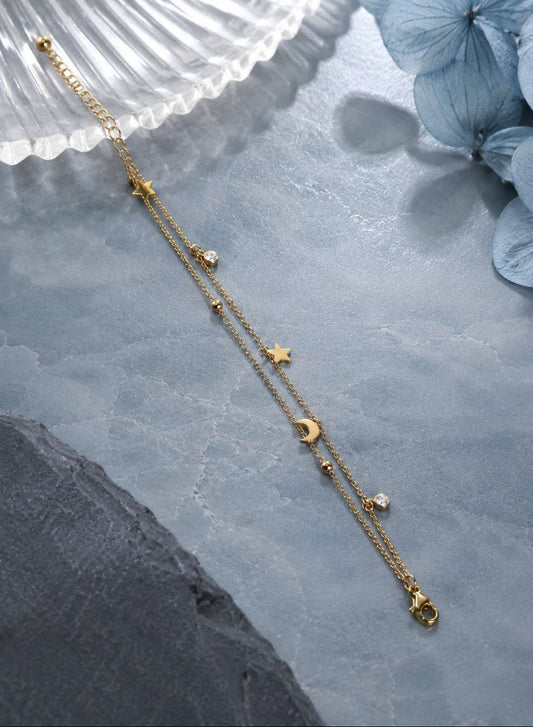 A delicate gold-toned double-strand chain necklace with celestial charms, including stars, moons, and small beads, styled on a textured gray surface with a white shell and blue flowers.