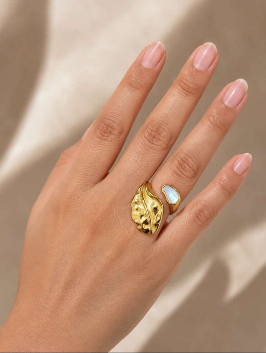 A close-up of a hand wearing a gold leaf-shaped ring with a white opal inlay, set against a warm beige background with soft shadows.