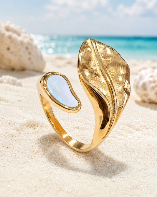 A gold open ring with a large textured leaf-like side and a smooth oval white inlay, photographed standing in sand with a beach and ocean blurred in the background.