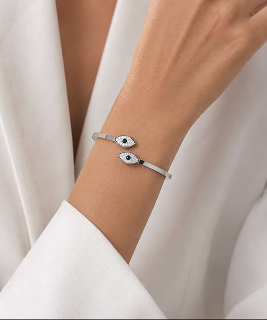 A close-up of a person's wrist wearing a slim silver cuff bracelet with two evil eye charms, styled with a white draped garment.