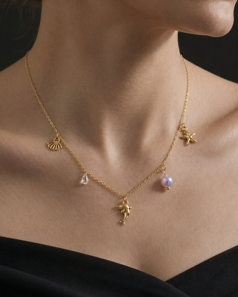 A close-up of a model wearing a delicate gold necklace with small charms, including a pearl bead and a teardrop-shaped stone, against a dark background.