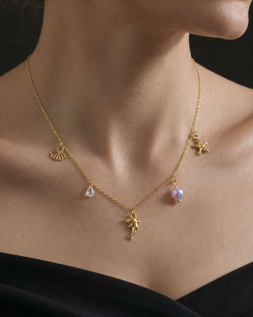 A close-up of a model wearing a delicate gold necklace with small charms, including a pearl bead and a teardrop-shaped stone, against a dark background.
