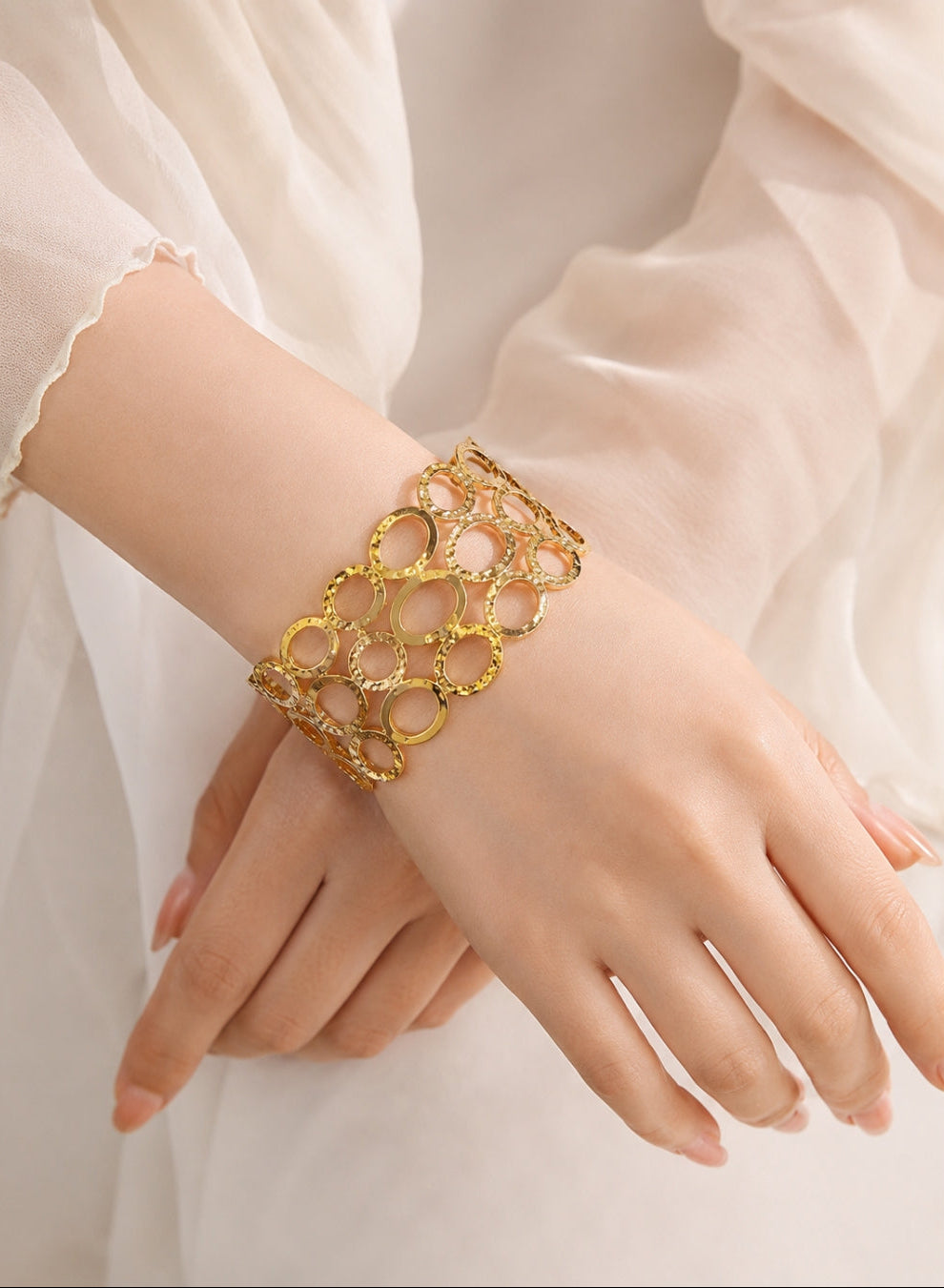 A gold-toned openwork circular link bracelet worn on a woman's wrist, shown against a soft cream-colored sheer dress fabric.