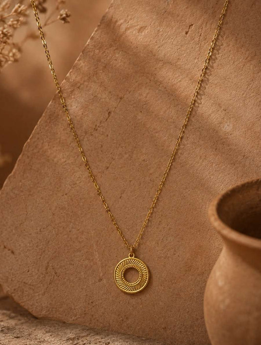 A gold necklace with a circular pendant featuring concentric textured rings, displayed on a rustic tan stone slab with dried flowers and a terracotta pot in the background.
