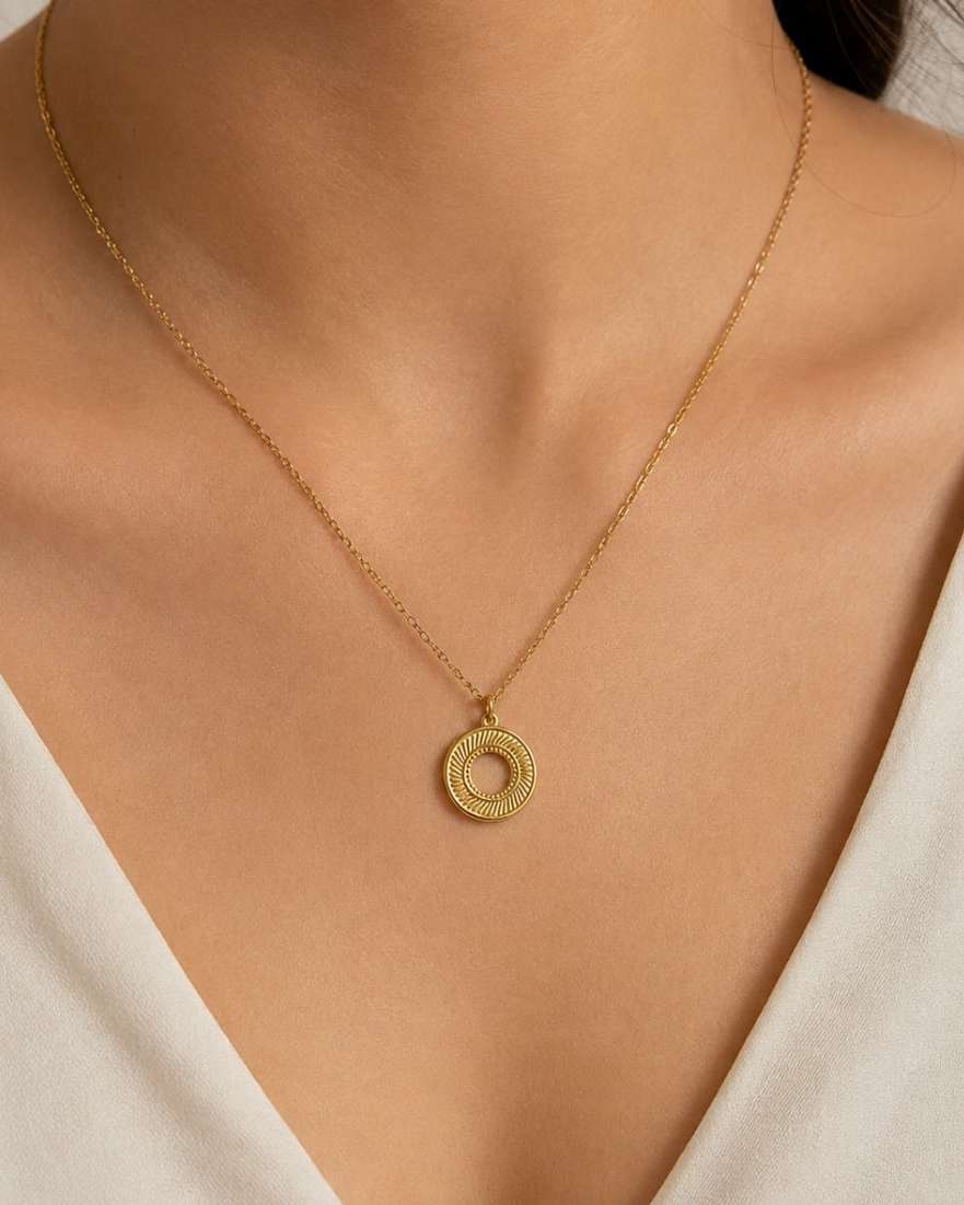 A close-up of a model wearing a delicate gold chain necklace with a textured circular ring pendant, styled with a cream-colored V-neck top.