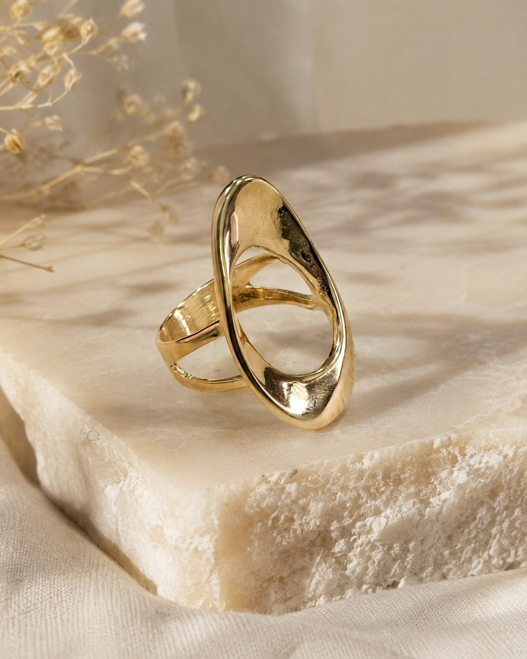 A polished gold abstract ring with a teardrop-shaped open design, resting on a textured cream-colored stone slab with dried floral elements in the background.