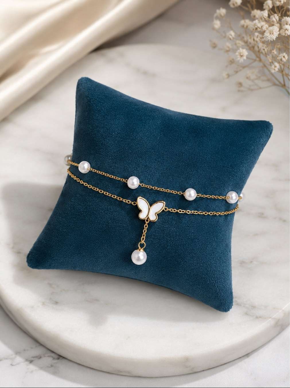 A double-strand gold chain bracelet with white pearl accents and a butterfly charm, displayed on a dark teal velvet pillow over a white marble surface.