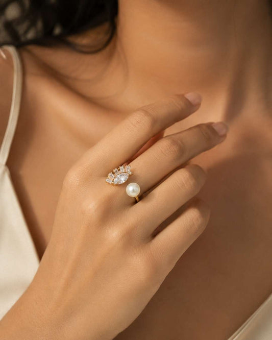 A close-up lifestyle shot of a woman's hand wearing a delicate gold ring with a single white pearl and a small cluster of clear stones above it.