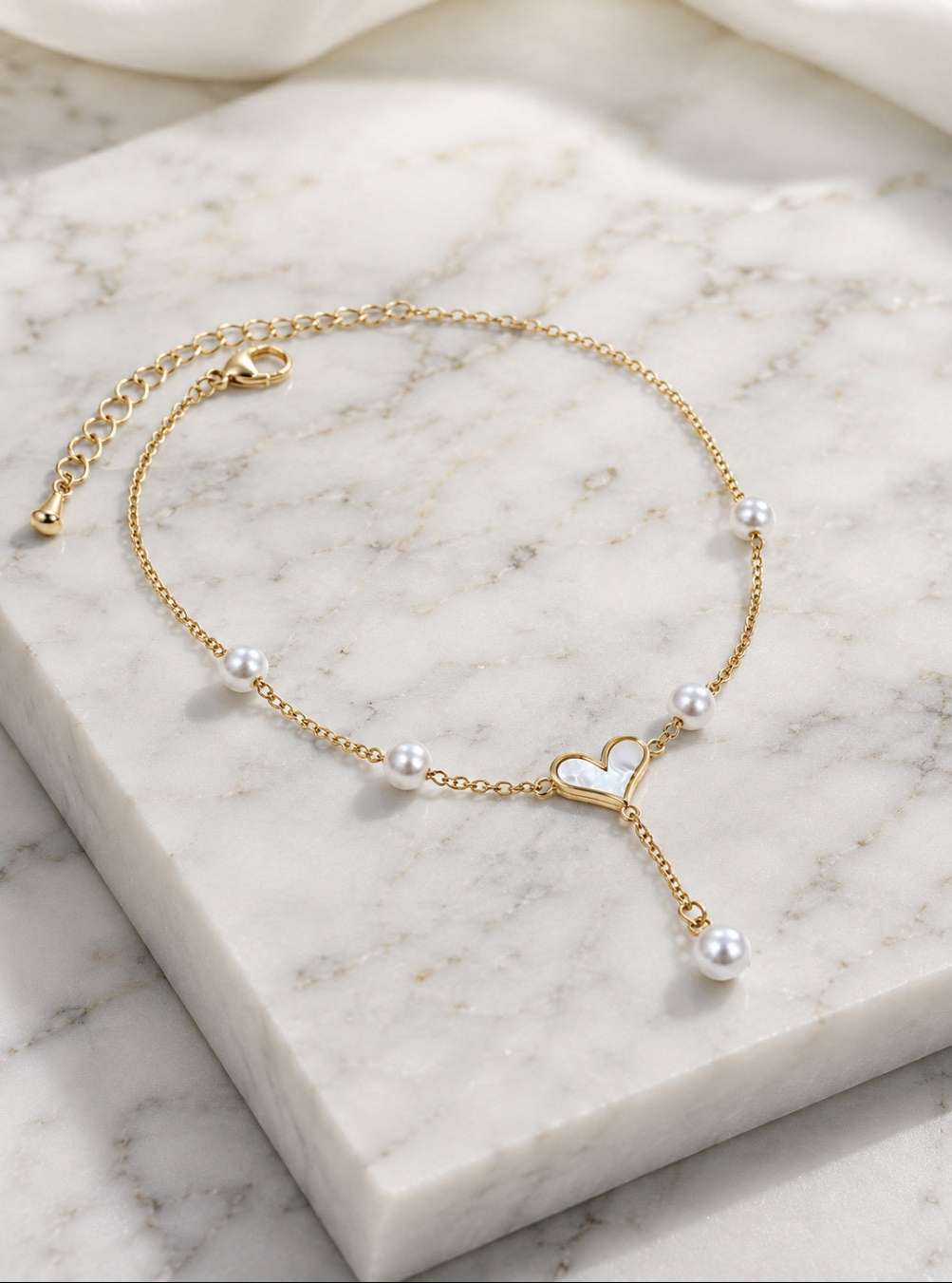 A delicate gold chain anklet with small white pearls and a central open heart charm, displayed on a white marble block.