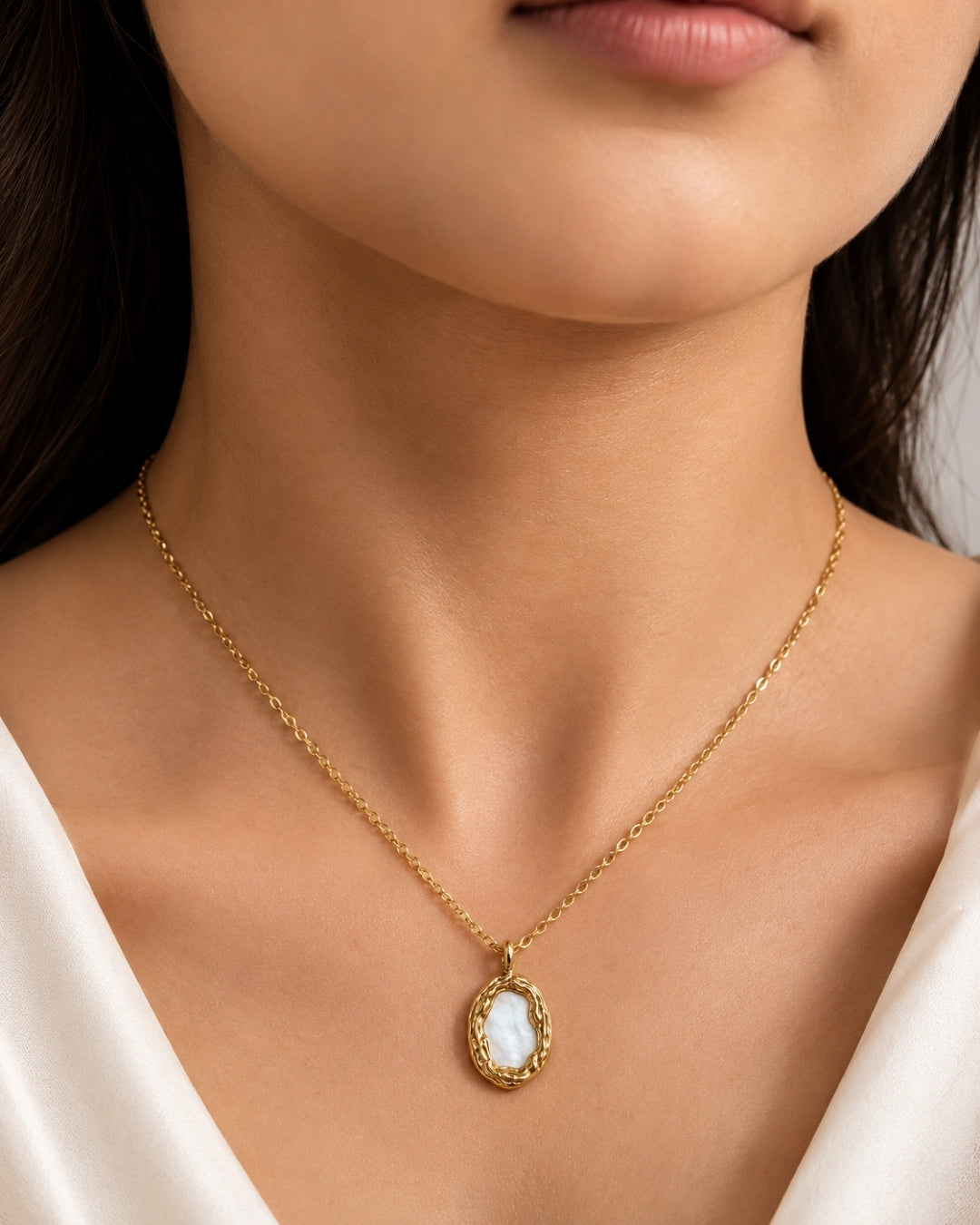 A close-up of a model wearing a delicate gold chain necklace with an oval pendant featuring a white mother-of-pearl pattern and a textured gold border.