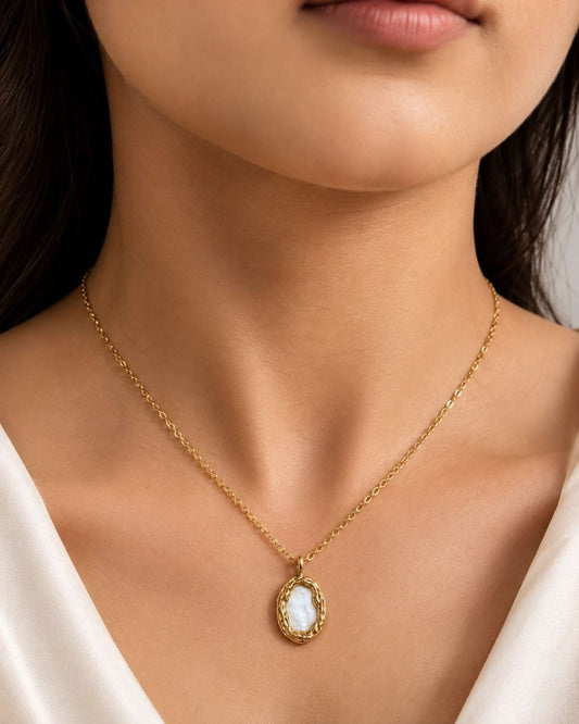 A close-up of a model wearing a delicate gold chain necklace with an oval pendant featuring a white mother-of-pearl pattern and a textured gold border.