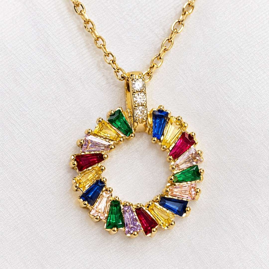 A gold chain necklace with a circular pendant set with colorful baguette gemstones in yellow, blue, green, pink, and red, displayed on a textured white fabric background.