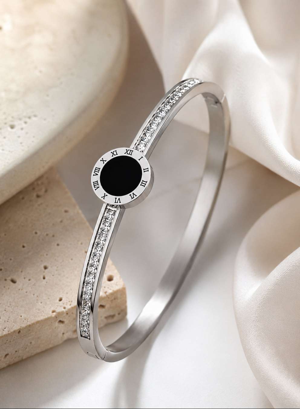 A polished silver-toned bangle bracelet with a central round black inlay and Roman numeral markers, accented with small clear stones along the band, styled on a white satin cloth with a beige stone prop.
