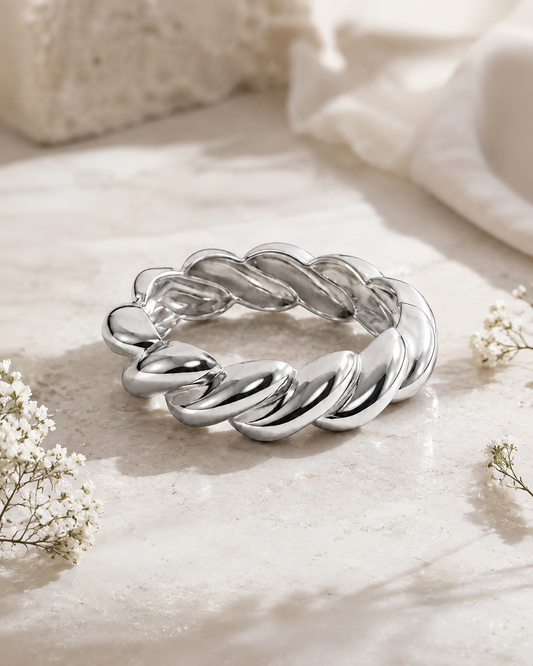 A polished silver twisted rope ring bracelet displayed on a light beige stone surface with soft white fabric and small white flowers in the background.