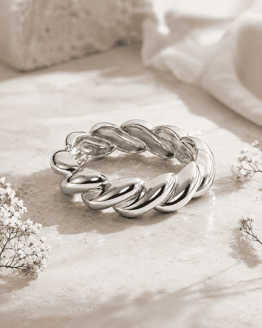 A polished silver twisted rope ring bracelet displayed on a light beige stone surface with soft white fabric and small white flowers in the background.