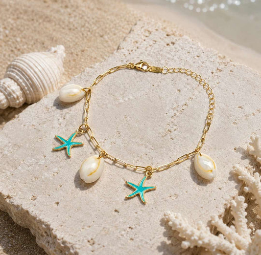 A gold chain bracelet with three white cowrie shells and two turquoise starfish charms, displayed on a textured stone with seashells and sand in the background.