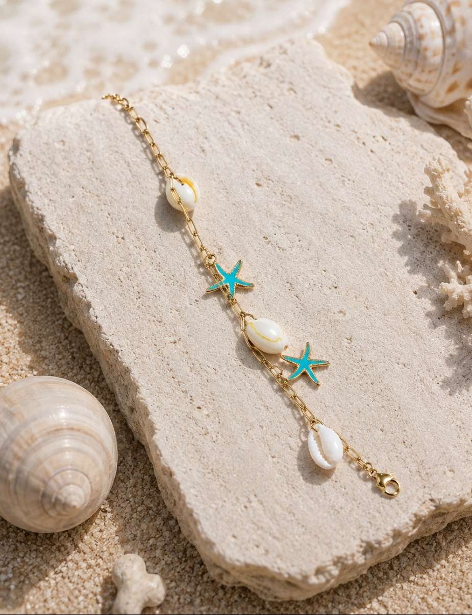 A gold chain bracelet with white seashells and turquoise starfish charms, displayed on a textured stone block over a sandy beach setting with real shells and coral.