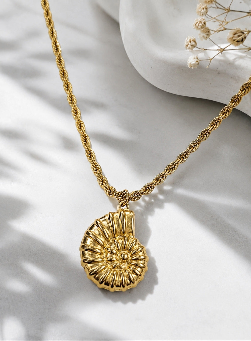 A gold necklace with a textured, shell-like pendant, displayed on a white surface with soft shadows and dried flowers in the background.