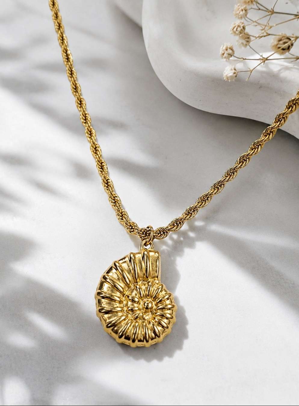 A gold necklace with a textured, shell-like pendant, displayed on a white surface with soft shadows and dried flowers in the background.