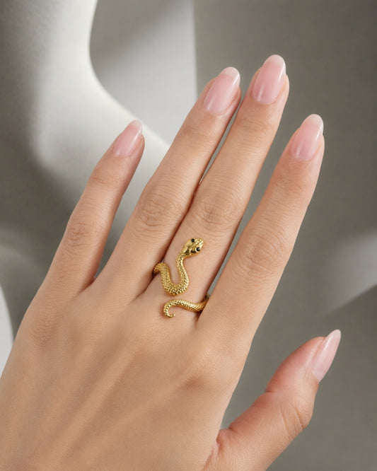 A close-up of a hand with long, neatly manicured pinkish nails wearing a small gold snake ring on the ring finger, set against a soft gray background.