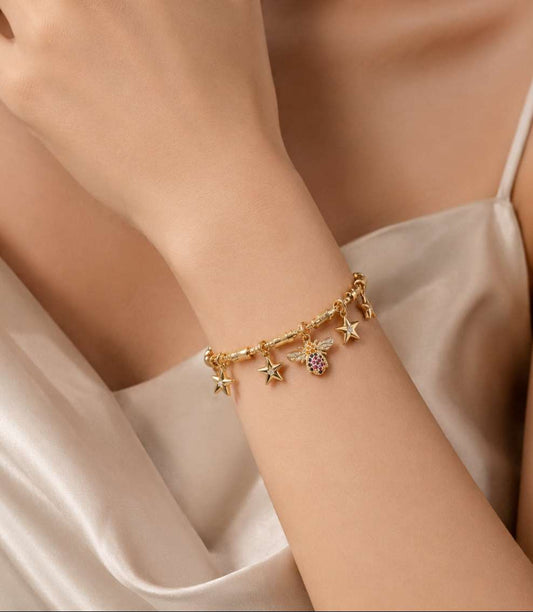 A gold-tone beaded anklet with purple and white beads, a central gold medallion featuring a turquoise and white mermaid design, and a lobster clasp, styled on white sand with seashells and a blue water background.