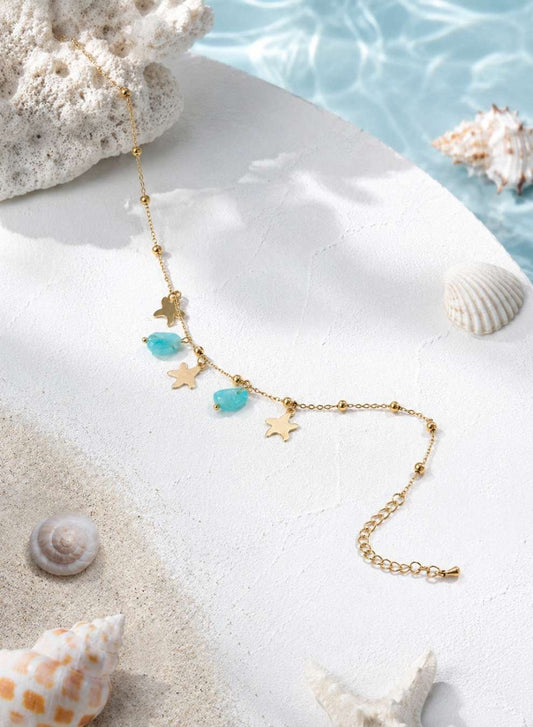 A gold-toned anklet with starfish and turquoise bead charms, styled on a white surface with seashells and a blue water backdrop.