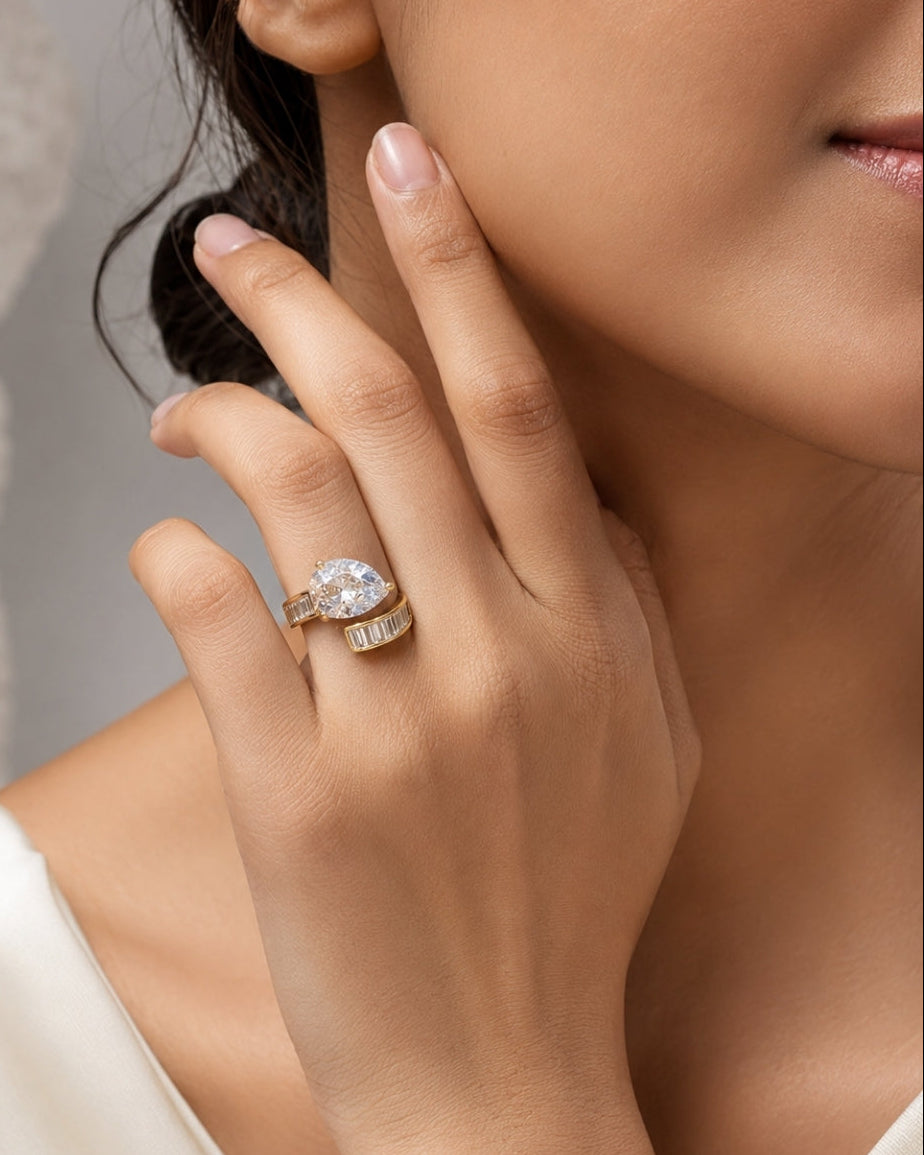 A close-up lifestyle shot of a woman wearing a gold ring with a large clear oval center stone and smaller side stones, resting her hand near her face.