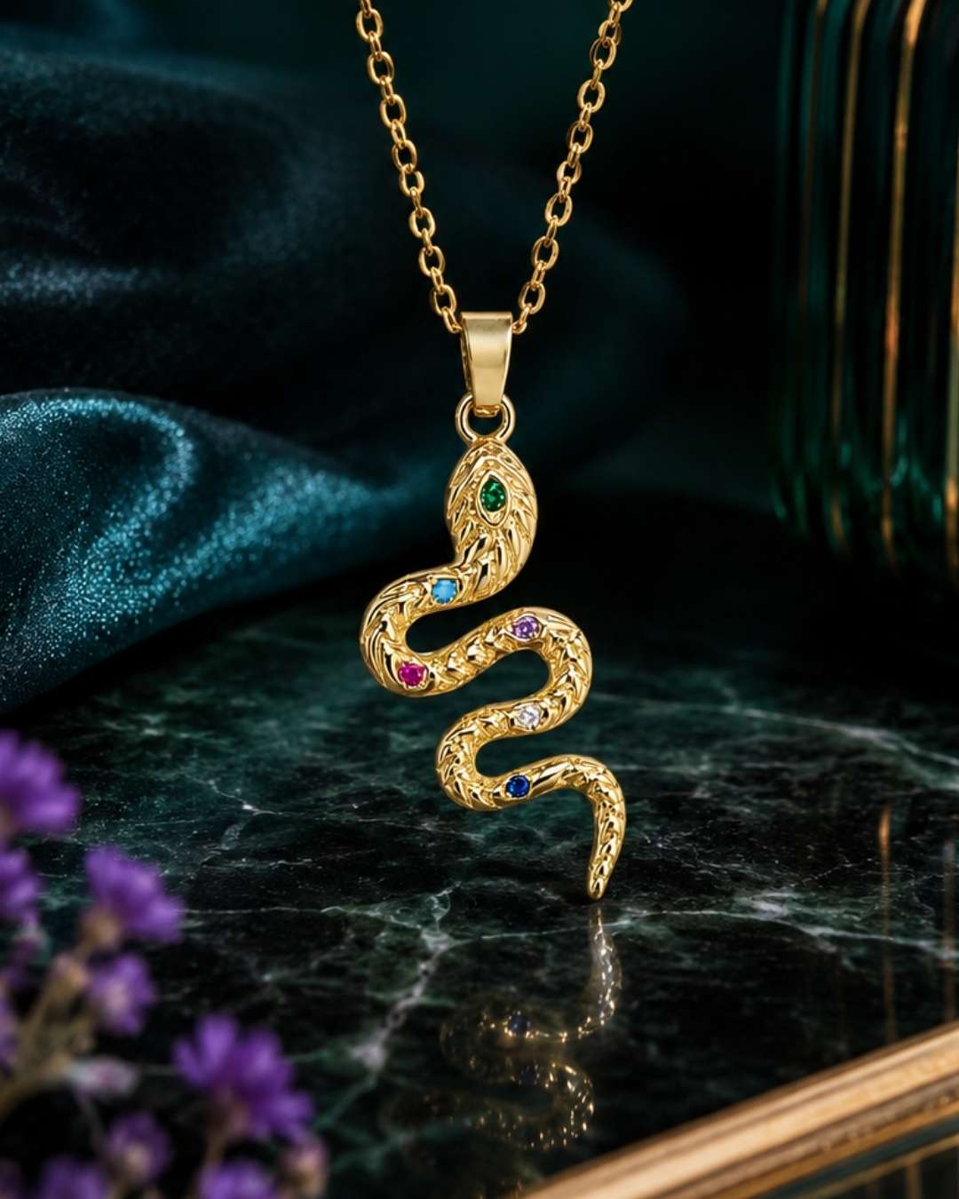 A gold snake pendant necklace with small multicolored gemstones, displayed on a dark marble surface with a teal satin cloth and purple flowers in the background.