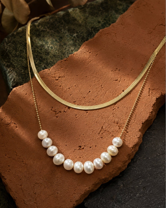 A layered necklace set displayed on a terracotta tile and dark stone background. The set includes a gold chain with a curved row of white freshwater pearls and a second gold chain with a smooth, curved gold pendant.